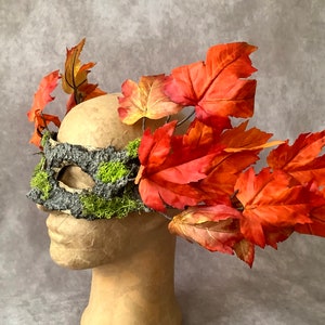 May include: A decorative mask featuring a bark-textured eye covering adorned with green moss and vibrant orange and red autumn leaves. The mask is designed to be worn, with the leaves extending around the head.