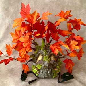May include: A handmade masquerade mask with a tree bark and moss design. The mask is adorned with a crown of orange and red fall leaves.