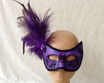 May include: Purple masquerade mask with black lace trim and rhinestones. A single purple feather is attached to the side.
