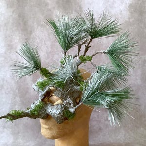 May include: A unique headpiece featuring a textured, gray and green base adorned with faux pine branches and needles. The branches are accented with white, creating a wintery or woodland theme. The headpiece is mounted on a mannequin head.