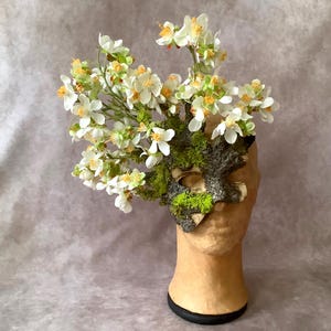 May include: A decorative mask with a natural, bark-like texture, adorned with white flowers and green moss. The mask is mounted on a mannequin head, with a black base. The flowers have yellow centers.