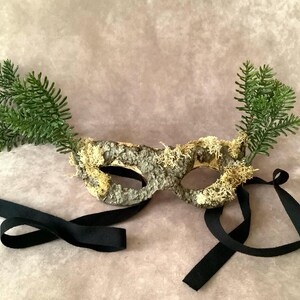 May include: A masquerade mask made of bark and moss with green sprigs attached to the sides. The mask is tied with a black ribbon.