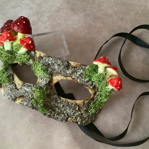 May include: A decorative mask with a woodland theme. The mask is adorned with red and white spotted mushrooms and green moss. The mask has a bark-like texture and is attached to a black ribbon.