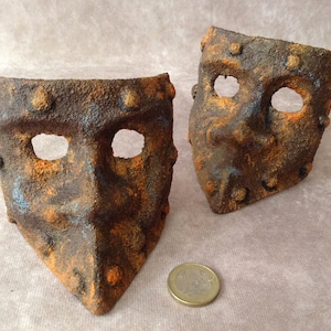 Può includere: Due maschere di metallo arrugginite con fori per gli occhi. Le maschere sono dipinte con vernice arancione, marrone e blu.