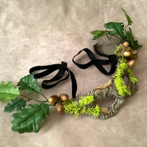 May include: A woodland-themed masquerade mask made from bark, moss, and acorns. The mask is attached to a black ribbon for wearing.