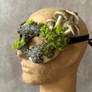 May include: A masquerade mask made of bark and moss with a cluster of mushrooms attached to the side. The mask is attached to a black elastic band.