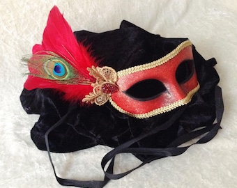 May include: A red masquerade mask with gold trim and a red feather with a peacock feather accent. The mask is on a black velvet cushion.
