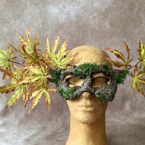 May include: A handmade mask made of bark and moss, with green moss accents and a pair of eyes. The mask is decorated with fall-colored maple leaves.