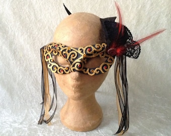 May include: A black masquerade mask with gold accents and red rhinestones. The mask has a delicate lace trim and a single red feather attached to the side.
