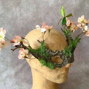 May include: A decorative mask with a woodland theme. The mask is adorned with faux pink flowers, green moss, and branches. The mask is placed on a mannequin head.