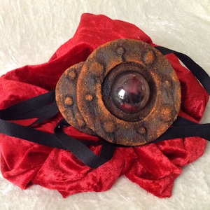 May include: A circular, rusty-brown, metal pendant with a black ribbon. The pendant has a circular, clear glass lens in the center.