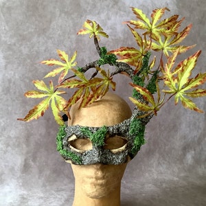 May include: A gray and green masquerade mask with moss and leaves, worn on a mannequin head with a leafy headdress.