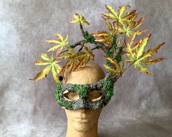 May include: A masquerade mask featuring a textured gray bark-like surface adorned with bright green moss. A branch extends from the top, decorated with several maple leaves in shades of yellow, light green, and reddish-brown, creating a forest-inspired look.