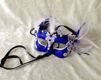 May include: A royal blue masquerade mask with white lace detailing and embellishments. The mask features white feathers and blue jewels. Black ribbons are attached for securing the mask.