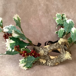 May include: A masquerade mask made of bark and moss with holly leaves and red berries attached. The mask has a black elastic band for wearing.