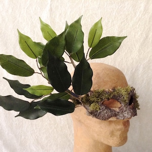 May include: A masquerade mask made of bark and moss with green leaves attached to the top. The mask is on a mannequin head.