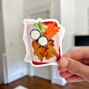 May include: A red and white basket filled with orange chicken nuggets, carrot sticks, celery sticks, and two white dipping sauces.