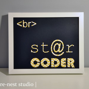 May include: A framed black chalkboard with white text that reads "<br> star@r CODER". The letters are in a white font with yellow chevron patterns. The frame is white.