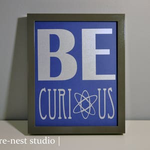 May include: A framed print with a blue background and white text that reads "BE CURIOUS" with an atom symbol in between the words.