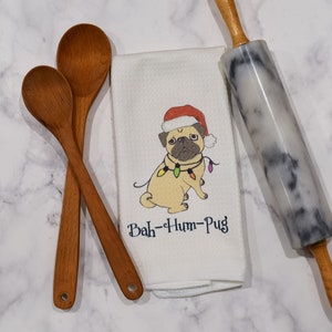May include: A kitchen scene with wooden spoons, a marble rolling pin, and a white dish towel. The towel features a pug wearing a Santa hat and Christmas lights, with the text "Bah-Hum-Pug".