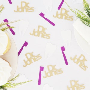 May include: White table with gold glitter confetti that says "Dr. Jones" and purple glitter confetti that says "Dr. Jones" and tooth and toothbrush shapes. There are also two cupcakes with white frosting on the table.
