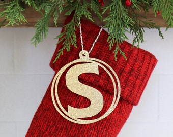 May include: A gold glittery circular ornament featuring the letter S in the center, suspended by a white and gold twisted cord. The ornament is positioned against a red knitted stocking with a ribbed cuff and green pine needles at the top.