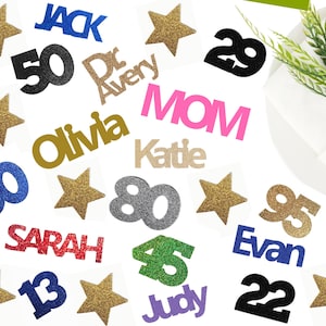 May include: Assorted glittery cutouts including gold stars, numbers, and names like "Jack," "Olivia," and "Evan." The cutouts are in various colors, such as blue, black, and pink, and are likely for party decorations.
