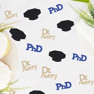 May include: White table with gold and blue confetti. The confetti features graduation caps, the text "Dr. Avery" and the text "PhD".