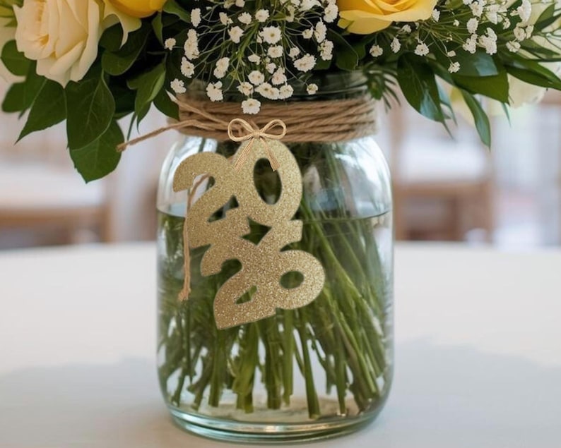 May include: A clear glass jar filled with water and green stems, holding a bouquet of yellow and white roses and baby's breath. A gold glitter '2020' ornament hangs from the jar, which is wrapped with twine.