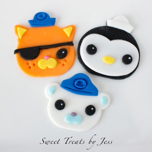 May include: Three colorful fondant decorations in the shape of cartoon characters. An orange cat with an eye patch, a black and white penguin, and a white bear, all wearing blue hats. The text "Sweet Treats by Jess" is at the bottom.