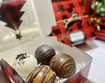Puede incluir: Una caja de cuatro esferas de chocolate gourmet, cada una con un diseño único. La caja tiene un patrón de cuadros rojos y negros. Una esfera es blanca con migas de galleta, otra es chocolate negro y dos son chocolate con leche con adornos decorativos.