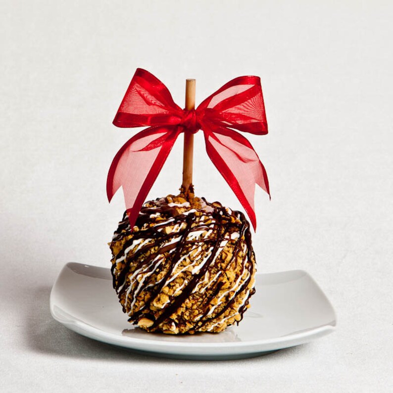 K&ouml;nnte beinhalten: Ein Schokoladen-Apfel mit Erdn&uuml;ssen und wei&szlig;em Schokoladen-Guss, gekr&ouml;nt mit einer roten Schleife auf einem wei&szlig;en Teller.