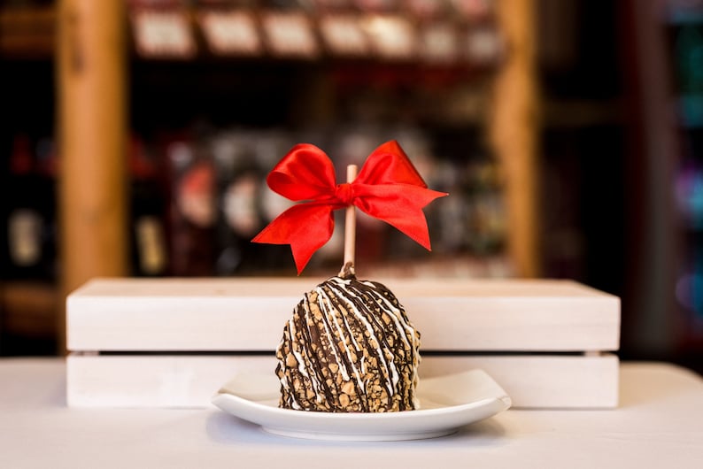 K&ouml;nnte beinhalten: Ein schokoladen&uuml;berzogener Apfel mit wei&szlig;en und braunen Bel&auml;gen ist mit einer roten Schleife verziert. Der Apfel liegt auf einem wei&szlig;en Teller und einer Holzkiste.