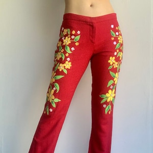 Op de afbeelding: Rode broek met bloemenborduurwerk. De broek heeft een rechte pijp en is versierd met gele en groene bloempatronen. De stof heeft een getextureerde uitstraling en de stijl is vintage.