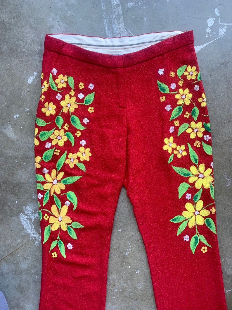 Op de afbeelding: Rode broek met een bloemenmotief. De broek heeft gele bloemen met groene bladeren en kleine witte bloemen. Het bloemenpatroon staat aan beide kanten. De broek heeft een rechte pijp.