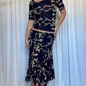 May include: A two-piece set featuring a short-sleeved top and a midi skirt. The set is navy blue with a tan abstract pattern. The top has a scoop neckline and the skirt has a ruffled hemline. The fabric appears to be sheer.