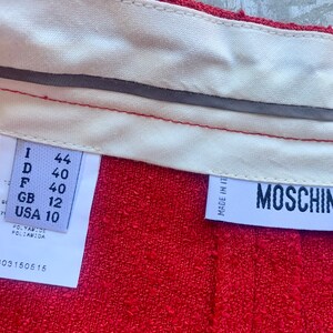 Op de afbeelding: Close-up van rode stof met een witte tailleband en labels. E&eacute;n label toont maatconversies: I 44, D 40, F 40, GB 12, USA 10. Het Moschino-label is ook zichtbaar. De stof is getextureerd.