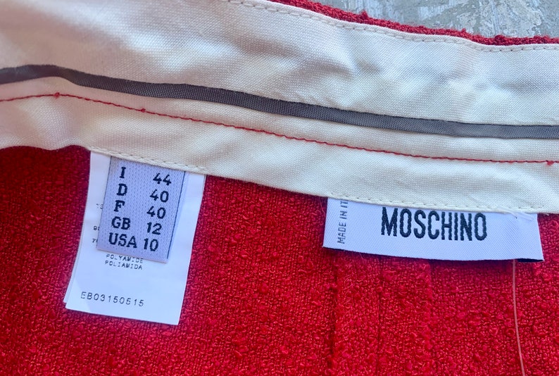 Op de afbeelding: Close-up van rode stof met een witte tailleband en labels. E&eacute;n label toont maatconversies: I 44, D 40, F 40, GB 12, USA 10. Het Moschino-label is ook zichtbaar. De stof is getextureerd.