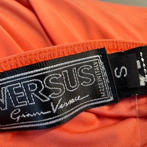 Może przedstawiać: Czarno-biała metka z tekstem "VERSUS Gianni Versace" i "MADE IN ITALY" w kolorze białym. Metka jest przymocowana do jasno pomarańczowego ubrania z metką rozmiaru "S".