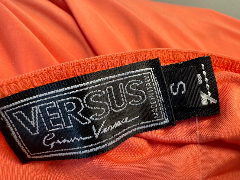 Może przedstawiać: Czarno-biała metka z tekstem "VERSUS Gianni Versace" i "MADE IN ITALY" w kolorze białym. Metka jest przymocowana do jasno pomarańczowego ubrania z metką rozmiaru "S".
