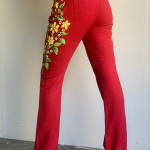 Op de afbeelding: Rode broek met bloemenborduurwerk. De broek heeft een textuurstof en een uitlopende pijp. Gele en groene geborduurde bloemen en bladeren vallen in een waterval langs het linkerbeen. De broek wordt gedragen met gele hakken.