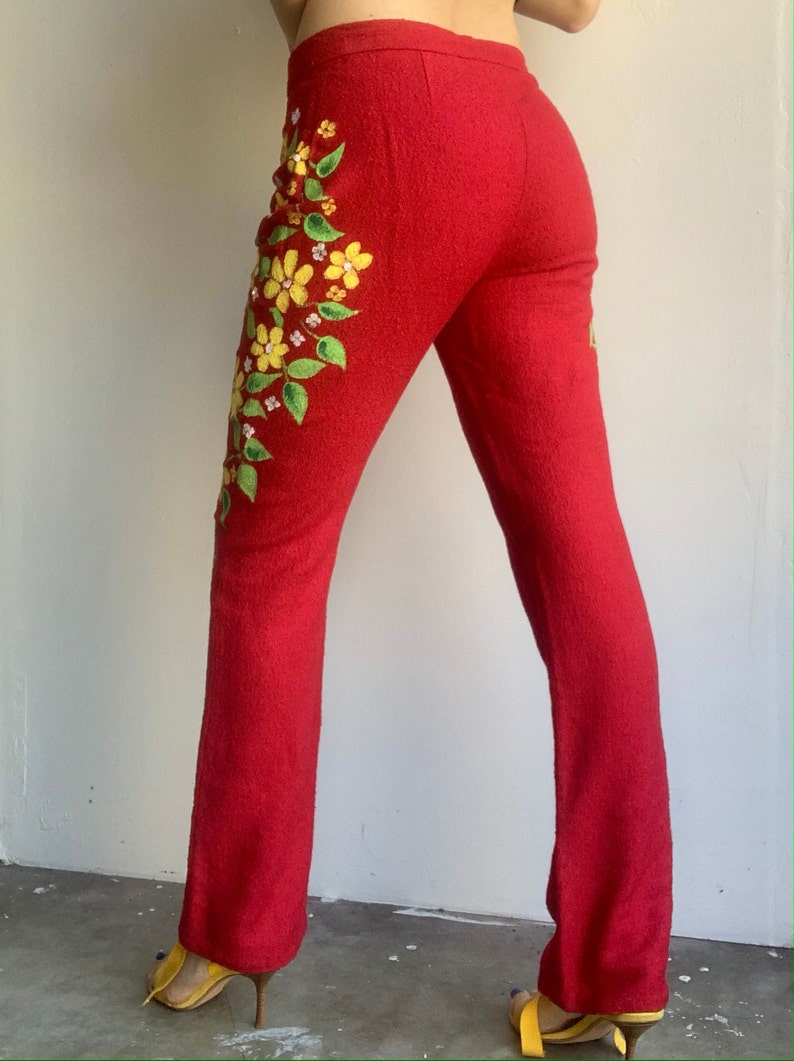 Op de afbeelding: Rode broek met bloemenborduurwerk. De broek heeft een textuurstof en een uitlopende pijp. Gele en groene geborduurde bloemen en bladeren vallen in een waterval langs het linkerbeen. De broek wordt gedragen met gele hakken.