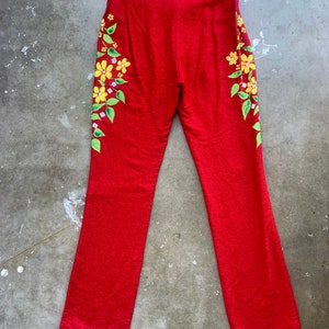 Op de afbeelding: Rode broek met een bloemenmotief. De broek heeft een levendige rode kleur en heeft een bloemenpatroon aan de zijkanten. De bloemen zijn geel met groene bladeren en kleine paarse accenten. De broek heeft een rechte pijp.