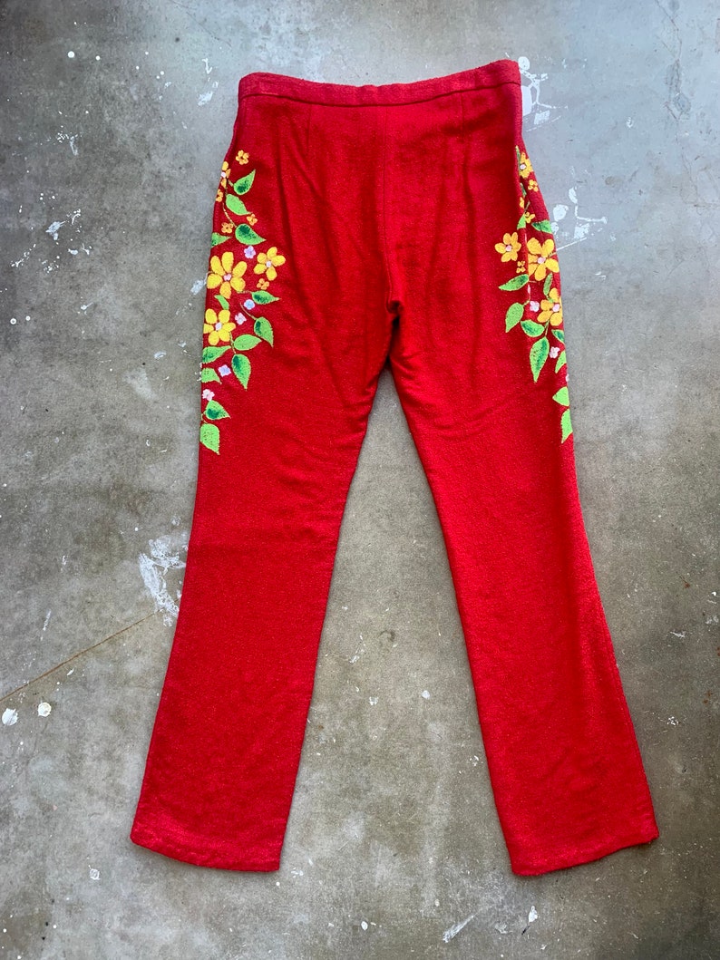 Op de afbeelding: Rode broek met een bloemenmotief. De broek heeft een levendige rode kleur en heeft een bloemenpatroon aan de zijkanten. De bloemen zijn geel met groene bladeren en kleine paarse accenten. De broek heeft een rechte pijp.