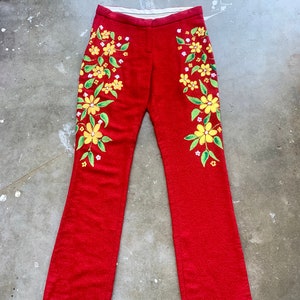 Op de afbeelding: Rode flared broek met gele bloemenborduurwerk. De bloemen hebben groene bladeren en witte accenten. De broek heeft een textuurstof en een witte tailleband. Een uniek, opvallend kledingstuk.