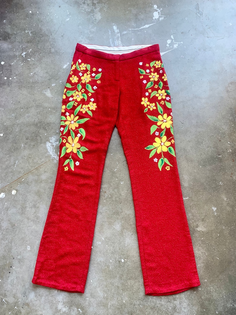 Op de afbeelding: Rode flared broek met gele bloemenborduurwerk. De bloemen hebben groene bladeren en witte accenten. De broek heeft een textuurstof en een witte tailleband. Een uniek, opvallend kledingstuk.