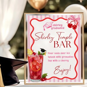 May include: A framed sign for a Shirley Temple Bar, featuring a pink and white striped background and red text. The sign includes a graduation cap graphic and a cocktail illustration. A black graduation cap with a gold tassel sits to the left.