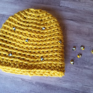 May include: A yellow crocheted beanie hat with a chunky knit design. The hat is adorned with small bee-shaped embellishments. Several bee embellishments are scattered nearby.