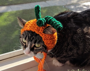 May include: A crocheted orange pumpkin hat for a cat, featuring a textured orange cap with a green stem and curly green vines on top. The hat includes two orange crocheted ties hanging down from the sides. The text To Craft a Home ©2018 is visible.