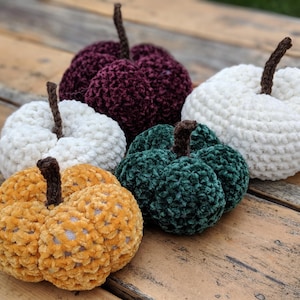 May include: Four crocheted pumpkins in different colours: burgundy, white, green, and yellow. The pumpkins are made with a soft, fuzzy yarn and have a textured surface.